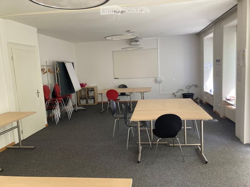 Meeting room with multiple tables, chairs, a projector screen, and a small shelf with storage bins.
