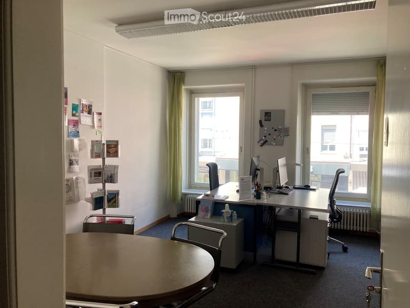 Office space with a desk, computer, chairs, a round table, and a filing cabinet. Windows with curtains provide natural light, and posters are displayed on the walls.