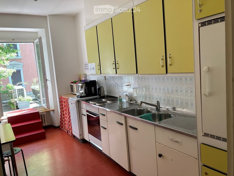 Bright kitchen with yellow upper cabinets, white lower cabinets, and a stainless steel sink. Appliances include a refrigerator, oven, microwave, and coffee maker. A small stool is near the window, and a red staircase leads outside.