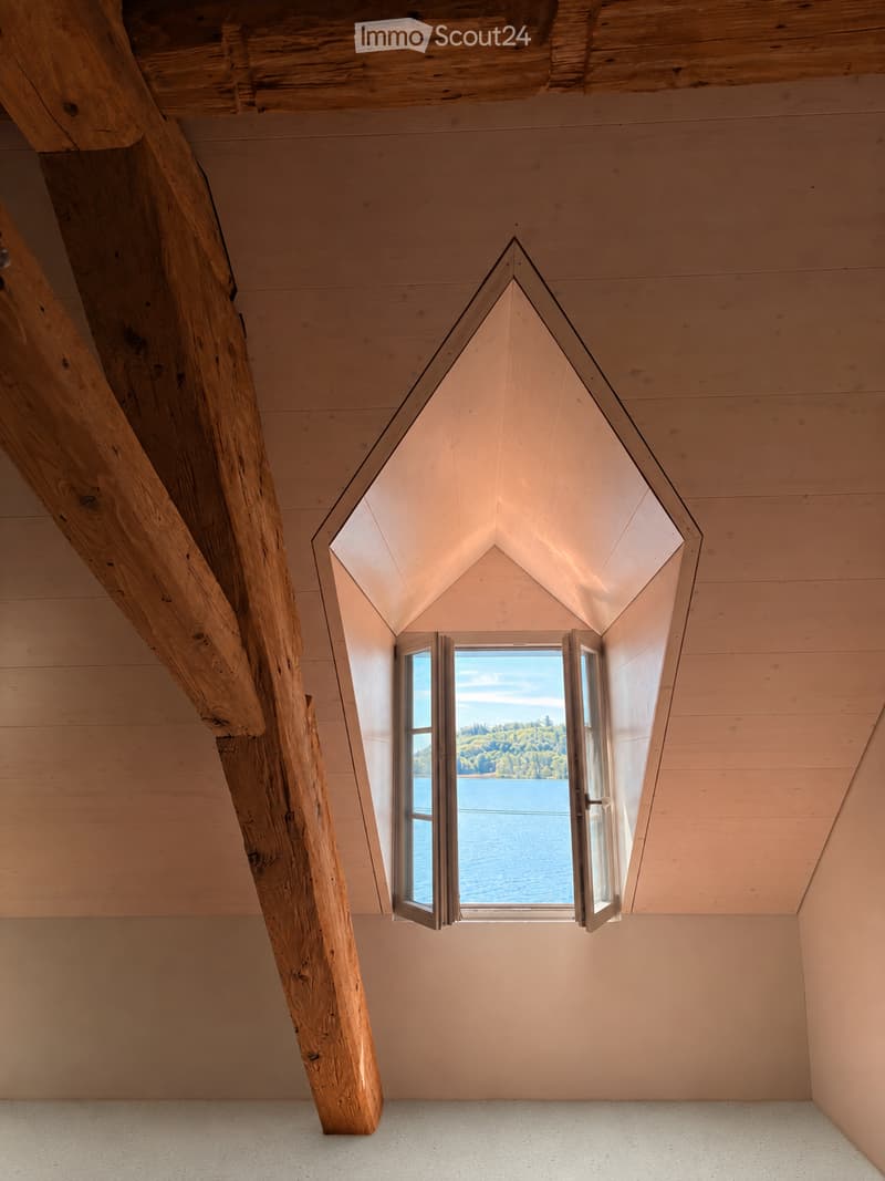 Unfurnished Room. An empty room with wooden beams and a large window overlooking a lake, featuring a sloped wooden ceiling and light-colored walls.