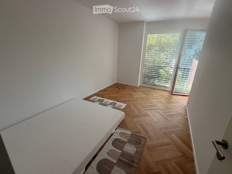 Bedroom. Bright bedroom with wooden herringbone flooring, white walls, and large windows with blinds. A single bed and a patterned rug are visible.