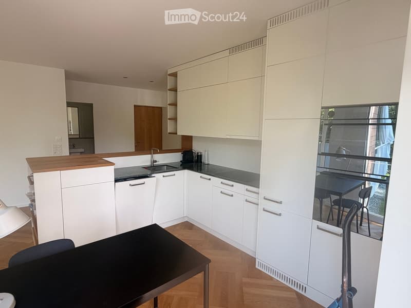 Furnished Room. Modern kitchen with white cabinets, wooden countertop, and a small dining area visible through a glass partition.