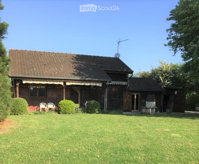 House. A two-story wooden house with a brown tiled roof, white-framed windows, and a covered porch area. The house is surrounded by a well-maintained lawn with shrubs and a few outdoor chairs.