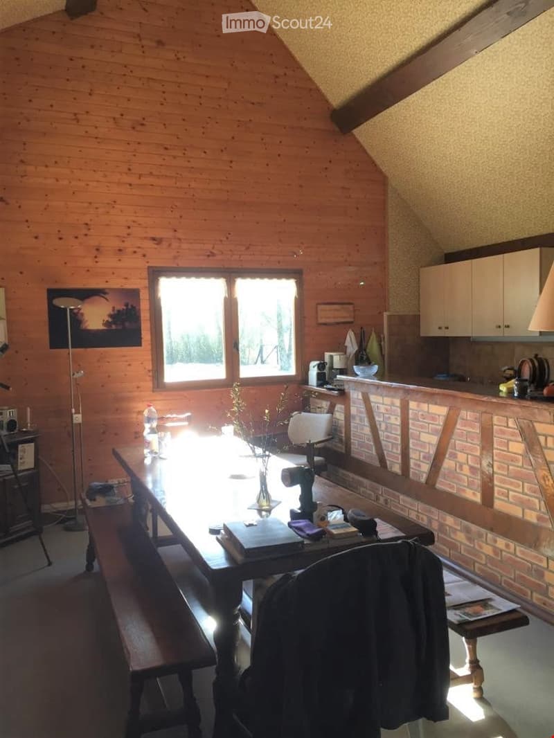 Furnished Room. A cozy attic room with wooden walls and a slanted ceiling. Features include a large wooden table with a bench, a chair, a cabinet, a lamp, a vase with flowers, a laptop, a bottle, a cup, and a fridge.