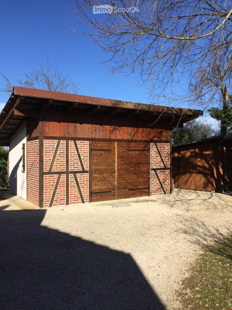 Building. A wooden garage with a brick facade and large wooden doors, situated on a gravel driveway under a clear blue sky.