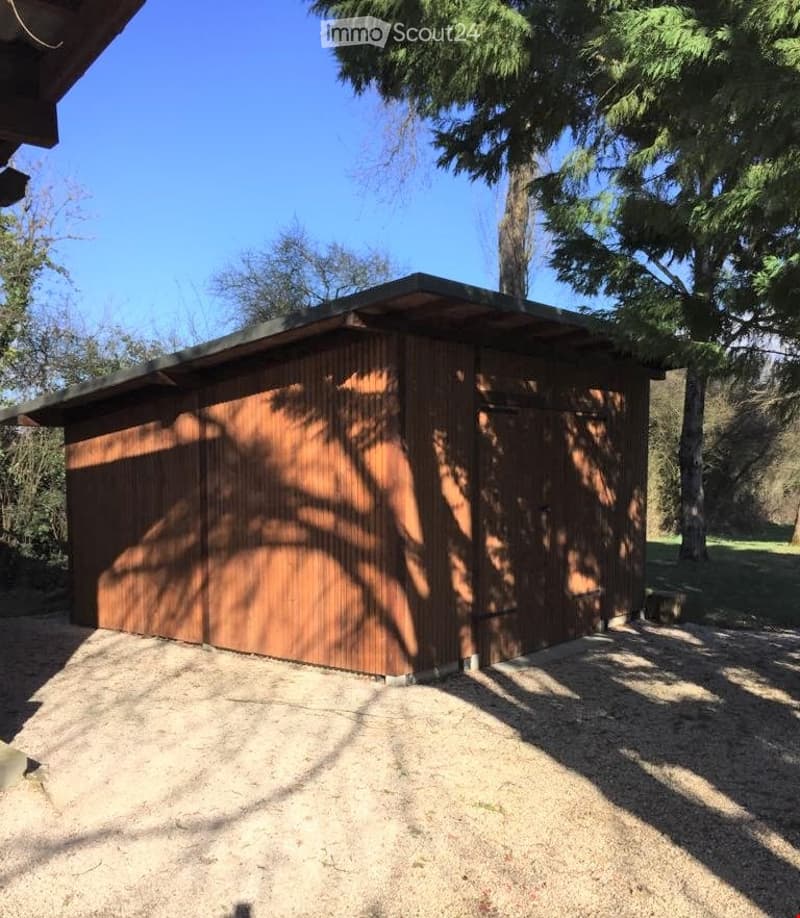 Building. A small wooden shed with a flat roof, situated in a garden area with trees and gravel ground.