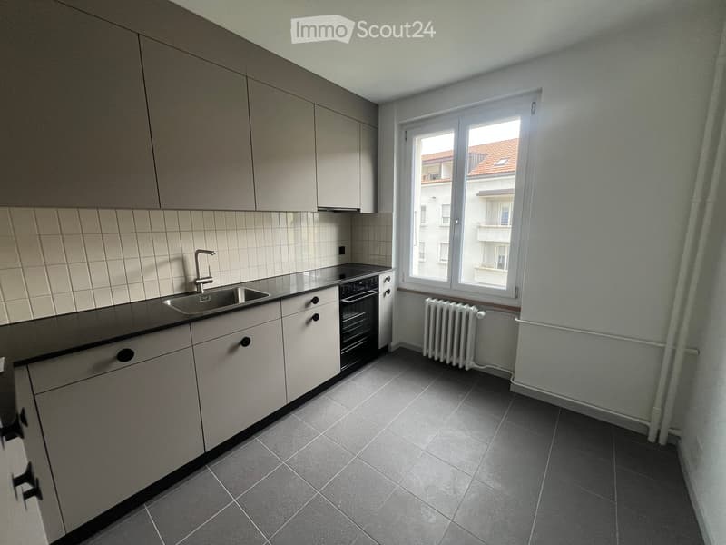 Modern kitchen with grey cabinets, black countertop, stainless steel sink, and built-in oven. Grey tiled floor and radiator under window.