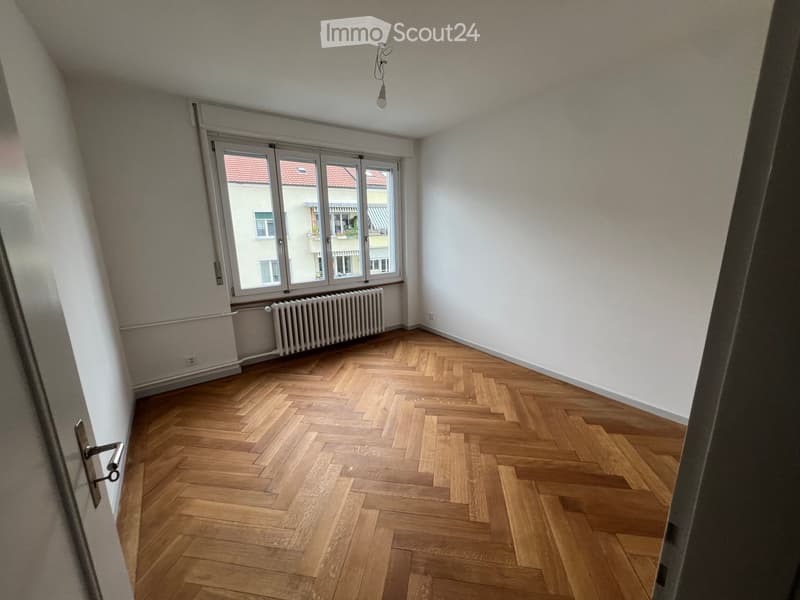 Empty room with wooden herringbone flooring, white walls, large window with view of neighboring building, radiator, and a single light fixture on the ceiling.