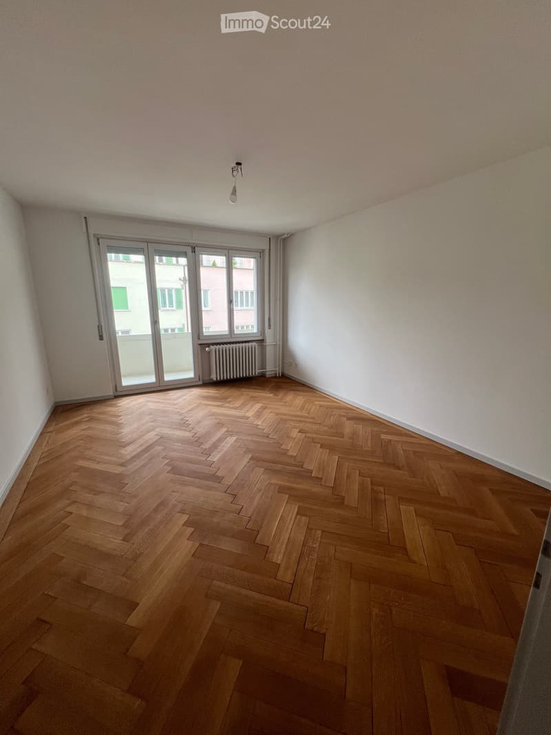 Empty room with wooden herringbone flooring, white walls, large sliding glass door, radiator, and ceiling light fixture.