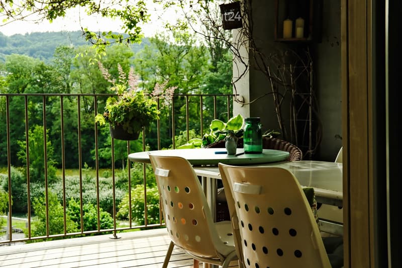 Balcony with a round table, two chairs, a vase, and potted plants, overlooking a lush green landscape.