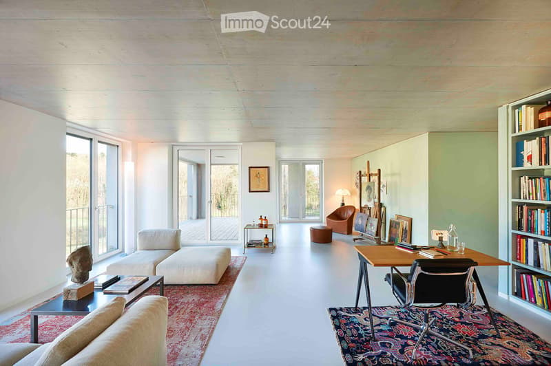 Furnished Room. Bright, open living space with white walls and ceiling, large windows, and sliding glass doors leading to a balcony. Features a white sectional sofa, a wooden desk with a black chair, a bookshelf, a small coffee table with a sculpture, and