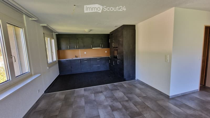 Modern kitchen with dark grey cabinets, a light orange backsplash, and a black tiled floor. The space includes a sink and faucet, with large windows allowing natural light to enter.