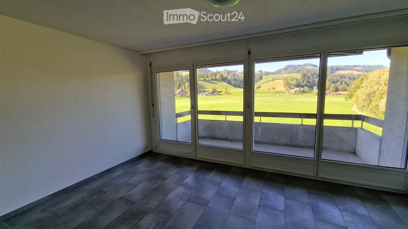 Empty room with large windows offering a scenic view of green fields and hills. The room has tiled flooring and white walls.