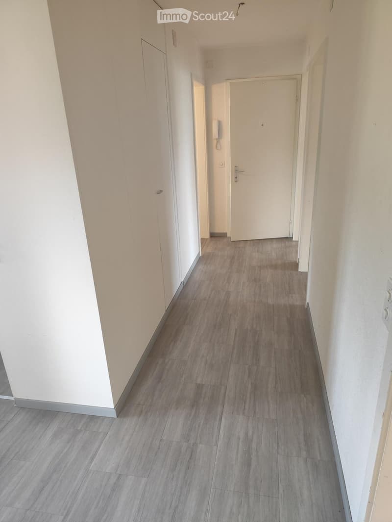 Empty hallway with light-colored walls and grey tiled flooring, featuring a closed white door at the end and a visible light switch on the right wall.