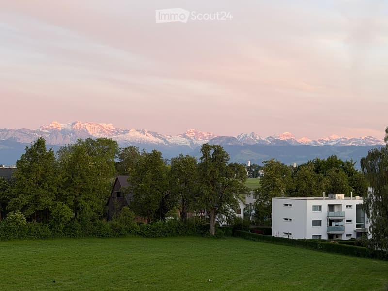 A modern white apartment building with balconies, surrounded by trees and a grassy lawn, with snow-capped mountains in the background.