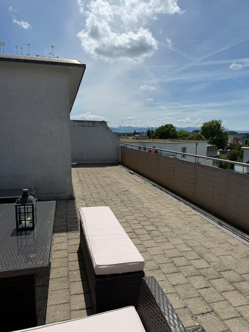 Roof terrace with brick tiles, a small table, two chairs, a bench, and a lantern, overlooking a cityscape with mountains in the background.