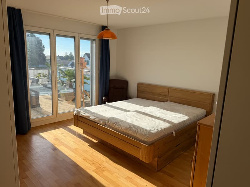 Bright bedroom with wooden floor, large window with sliding doors leading to a balcony, bed with white mattress, wooden headboard, and a single armchair.