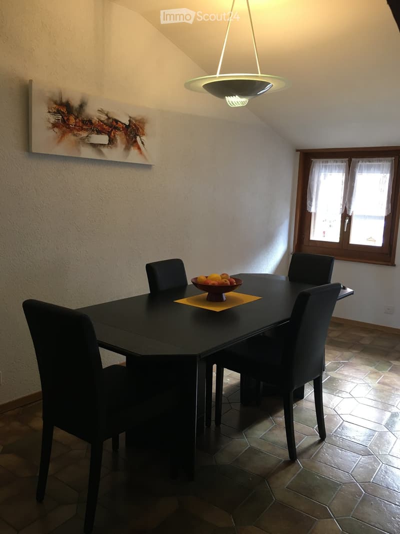 Dining room with a black dining table, four black chairs, a bowl of fruit on the table, a modern pendant light fixture, and a colorful abstract painting on the wall.