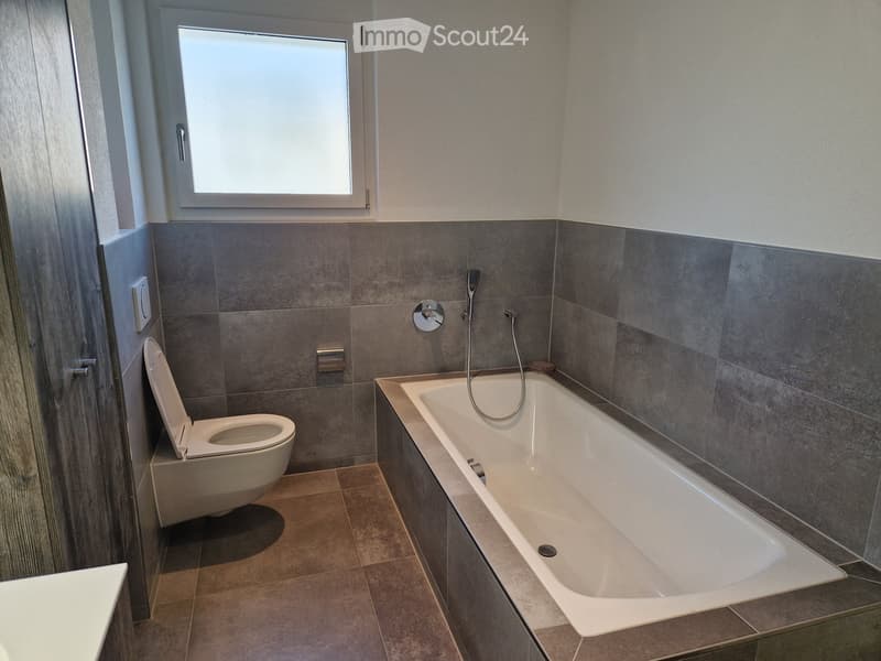 White bathtub, modern toilet, tiled walls, window, dark brown floor tiles