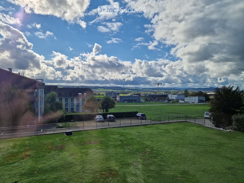 Aerial view of a grassy field with parked cars, a fence, several buildings in the background, and a cloudy sky.