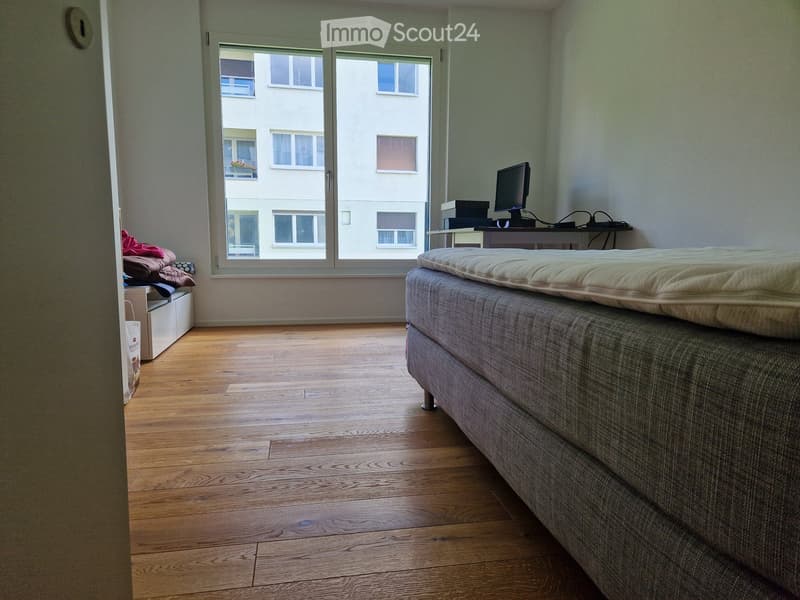 Bedroom with wooden floor, desk with monitor, large window, bed, clothes, and cabinets