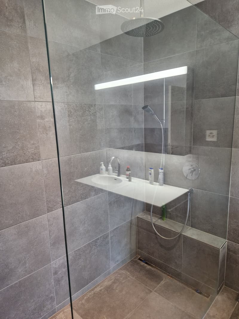 modern bathroom with glass shower enclosure, white sink, shower head, and shower hose, gray tiles, power outlet, and a bench