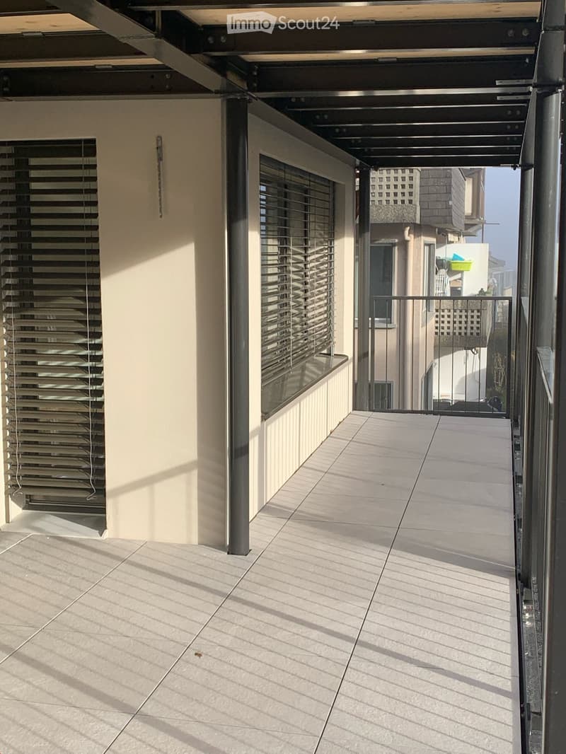 Modern balcony with tiled floor, metal railings, and large windows with blinds. The area is clean and well-lit with natural light.
