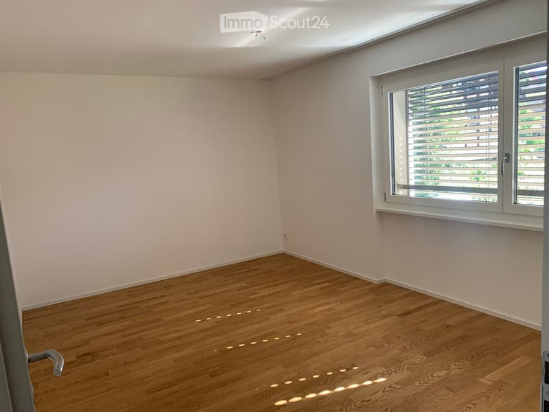 Empty room with light wooden flooring and white walls, featuring large windows with blinds and recessed ceiling lighting.
