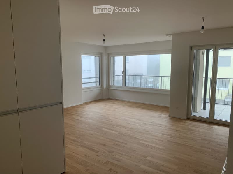 Empty room with wooden flooring, large windows, and sliding glass doors leading to a balcony.