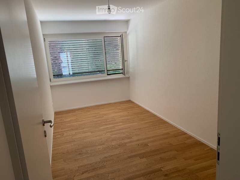 Empty room with wooden flooring, white walls, and a large window with blinds. The room is well-lit with natural light coming through the window.