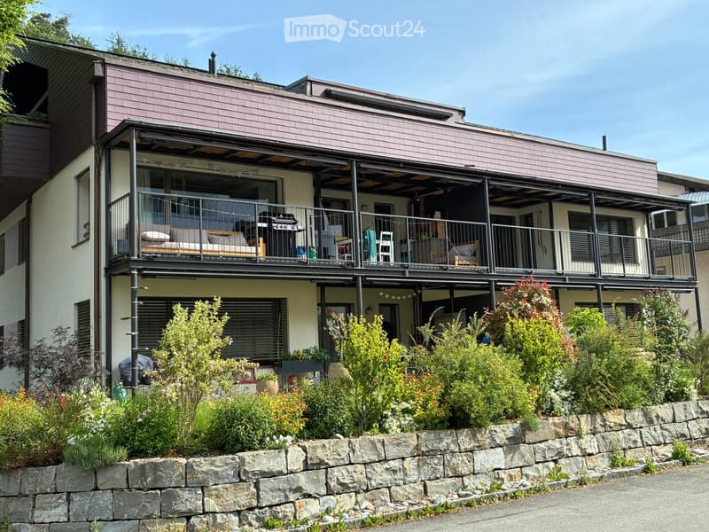 Modern two-story house with a large balcony, surrounded by greenery and a stone retaining wall.