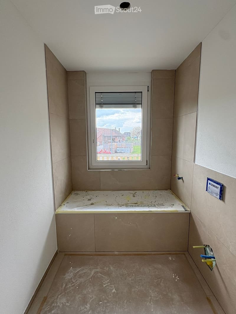 Empty bathroom with tiled walls, a window, and a bathtub base, showing construction materials and tools.