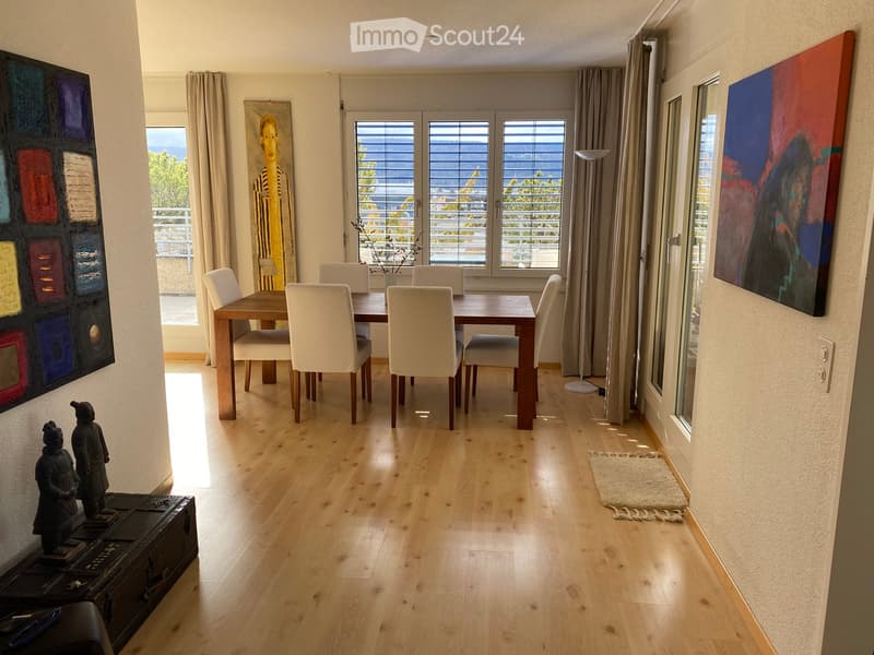 Bright dining area with wooden floor, large windows offering a scenic view, a dining table with six chairs, a floor lamp, and artwork on the walls.