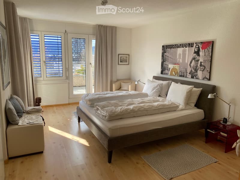 Modern bedroom with a large double bed, nightstand, chair, and artwork on the wall. The room features wooden flooring, large windows with blinds, and a cozy seating area.