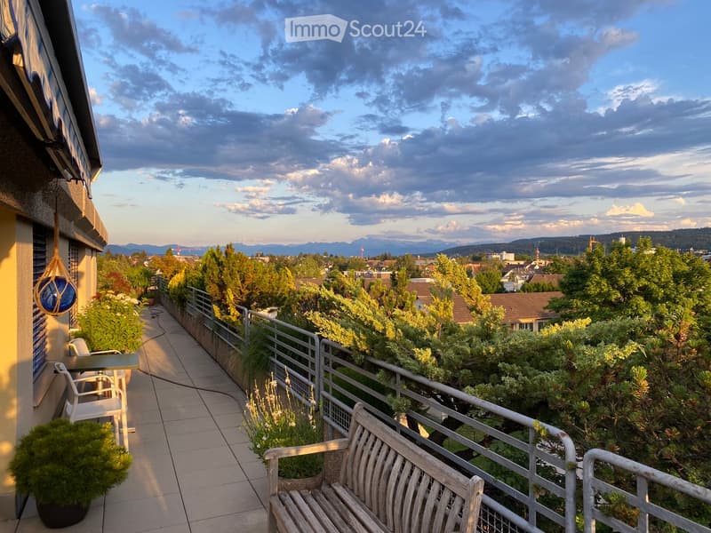 Balcony with tiled floor, metal railing, wooden bench, small table and chairs, potted plants, and a scenic city view.