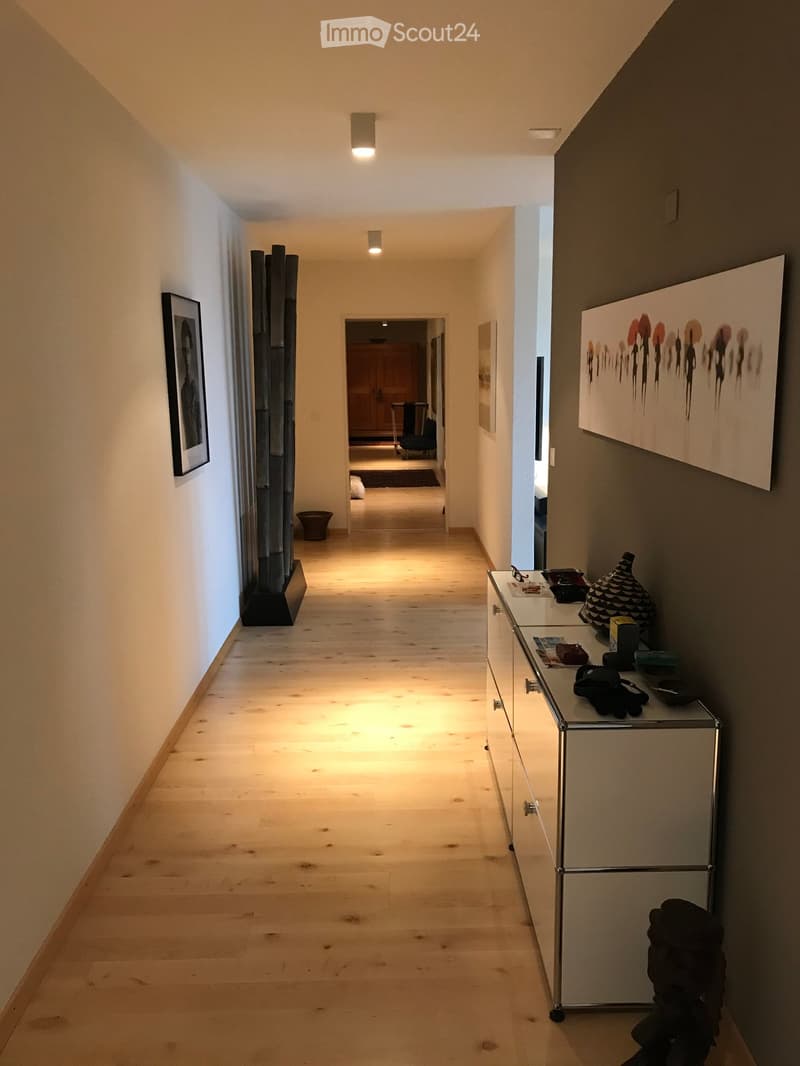 A long, well-lit hallway with wooden flooring, white walls, and modern decor including a glass console table, artwork, and a sculpture on the left side.