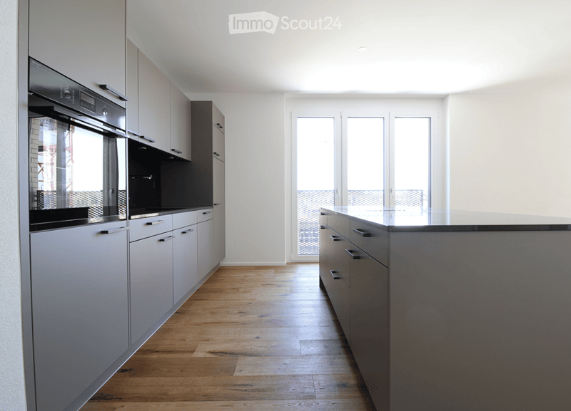 Modern kitchen with grey cabinets, wooden flooring, and an integrated oven. Large windows provide natural light.