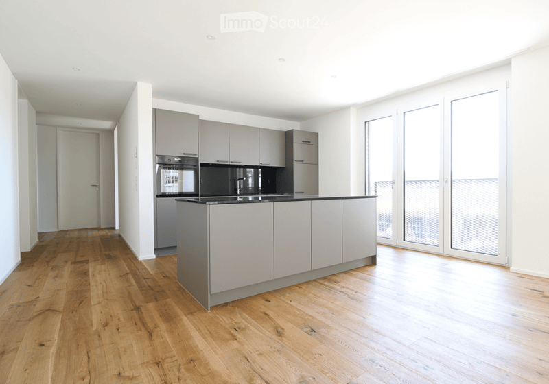 Modern kitchen with light grey cabinets, a central island, and a black countertop. The space features wooden flooring and large windows that allow natural light to fill the room.