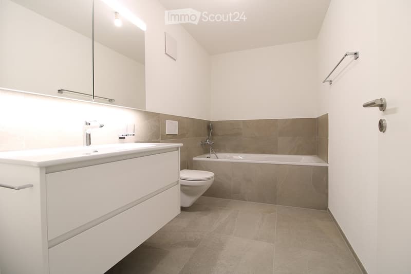 Modern bathroom with white cabinetry, a large sink, a bathtub, and a toilet. Features include a mirror, modern fixtures, and beige tiled walls and floor.