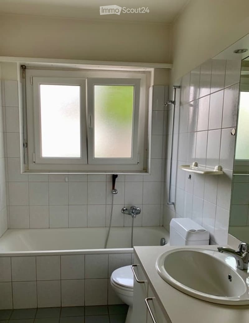Bathroom with white tiled walls and floor, a bathtub, toilet, sink, mirror, and a window with frosted glass panels.