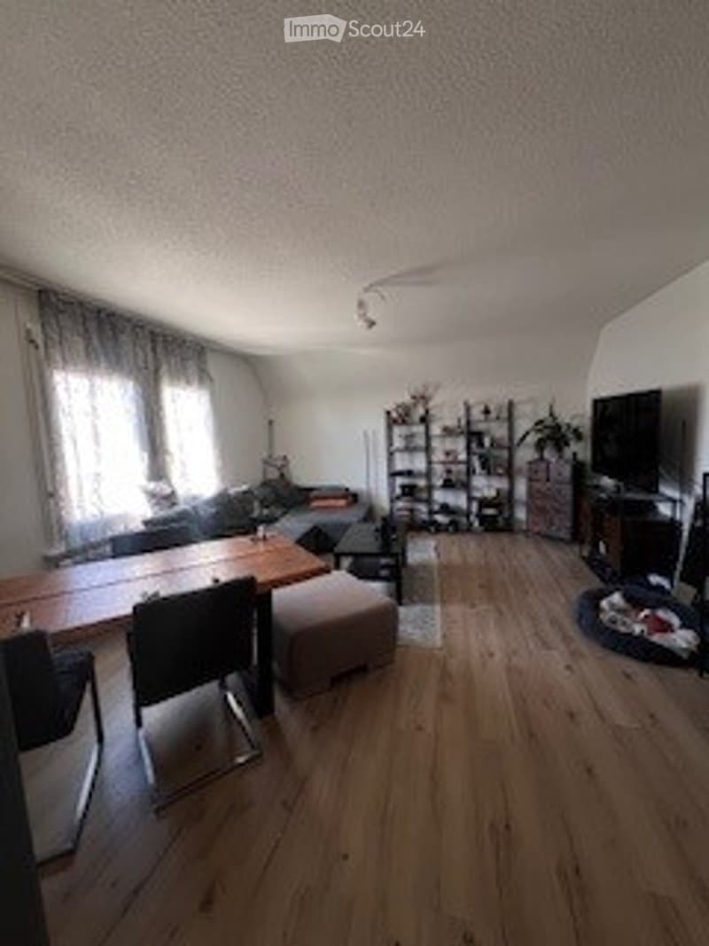 Bright living room with wooden flooring, large windows with curtains, a sofa, coffee table, ottoman, bookshelf, and television.