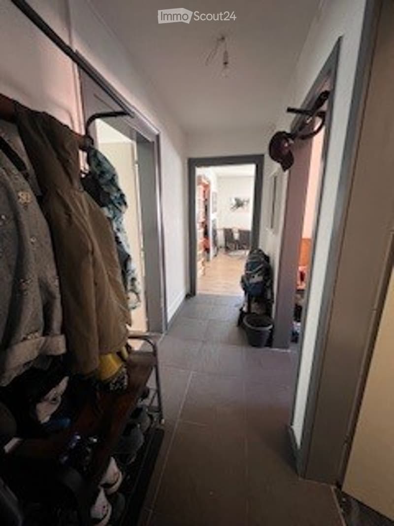 A narrow hallway with tiled flooring, coat rack on the left, and a small shoe rack. The hallway leads to an open doorway at the end, revealing part of another room.