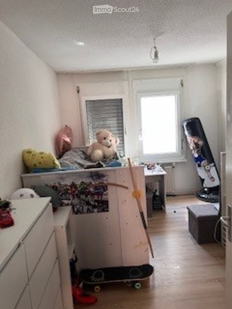 Small, furnished room with a bed, desk, chair, skateboard, teddy bear, and a stool. The room has wooden flooring and white walls.