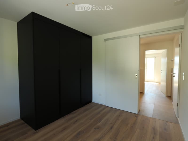 Unfurnished Room. Empty room with wooden flooring, white walls, and a large black wardrobe. An open doorway leads to another room with tiled flooring.