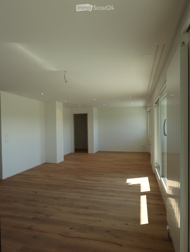 Unfurnished Room. Empty room with wooden flooring, white walls, and recessed lighting. Features a doorway leading to another space and large windows allowing natural light.