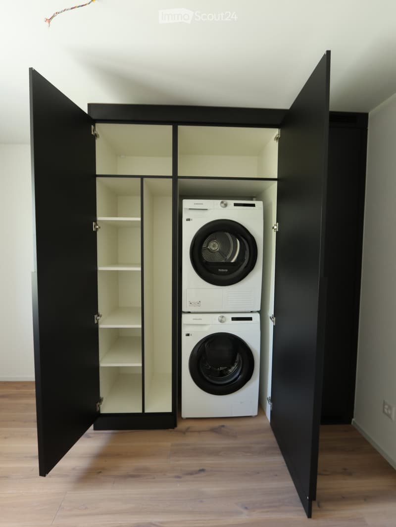 Furnished Room. A compact, modern laundry room with a stacked washing machine and dryer unit inside an open black cabinet with multiple shelves.