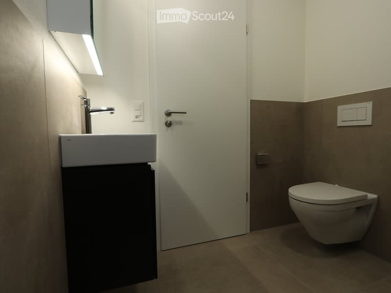 Modern bathroom with a white sink, chrome faucet, toilet, and built-in cabinet. Light-colored walls and tiled floor.