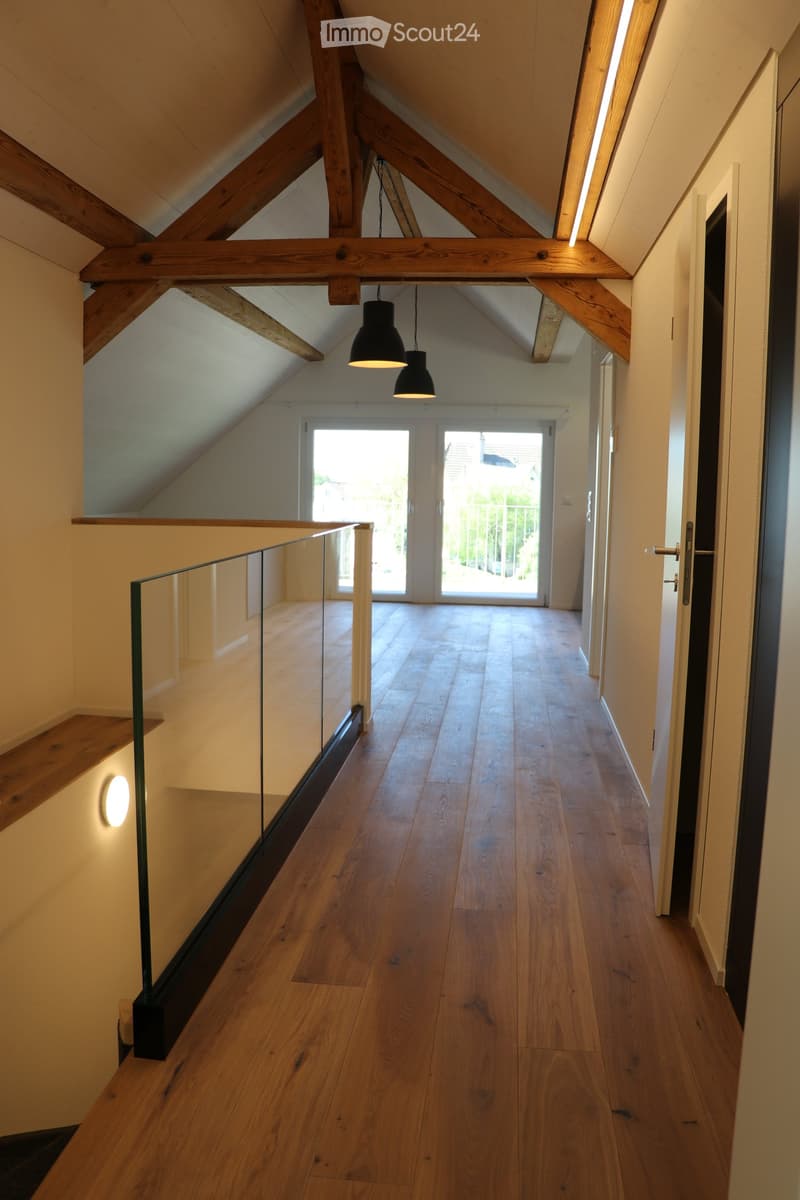 Spacious hallway with wooden flooring, exposed wooden beams, glass railing, and pendant lights.
