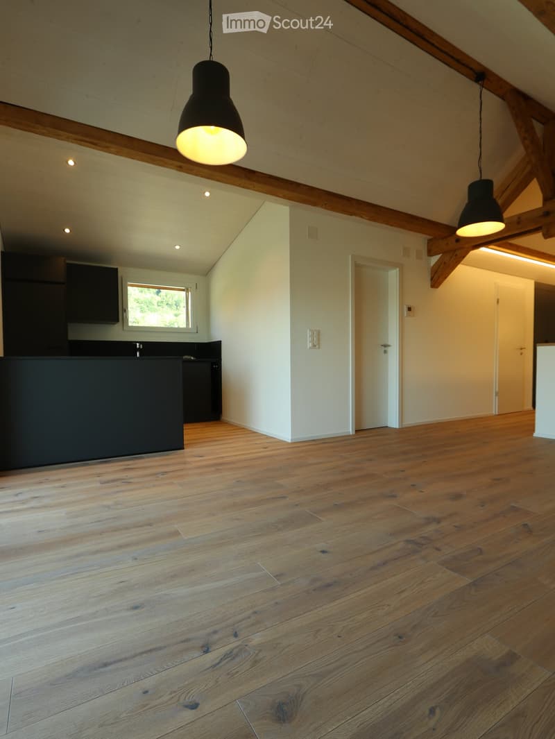Empty room with wooden flooring, white walls, exposed wooden beams, and pendant lights. Kitchen area visible in the background with cabinets and a countertop.