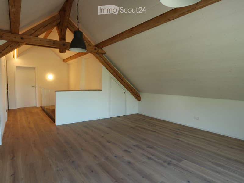 Empty attic room with wooden floors, exposed wooden beams, white walls, and a single hanging light fixture.